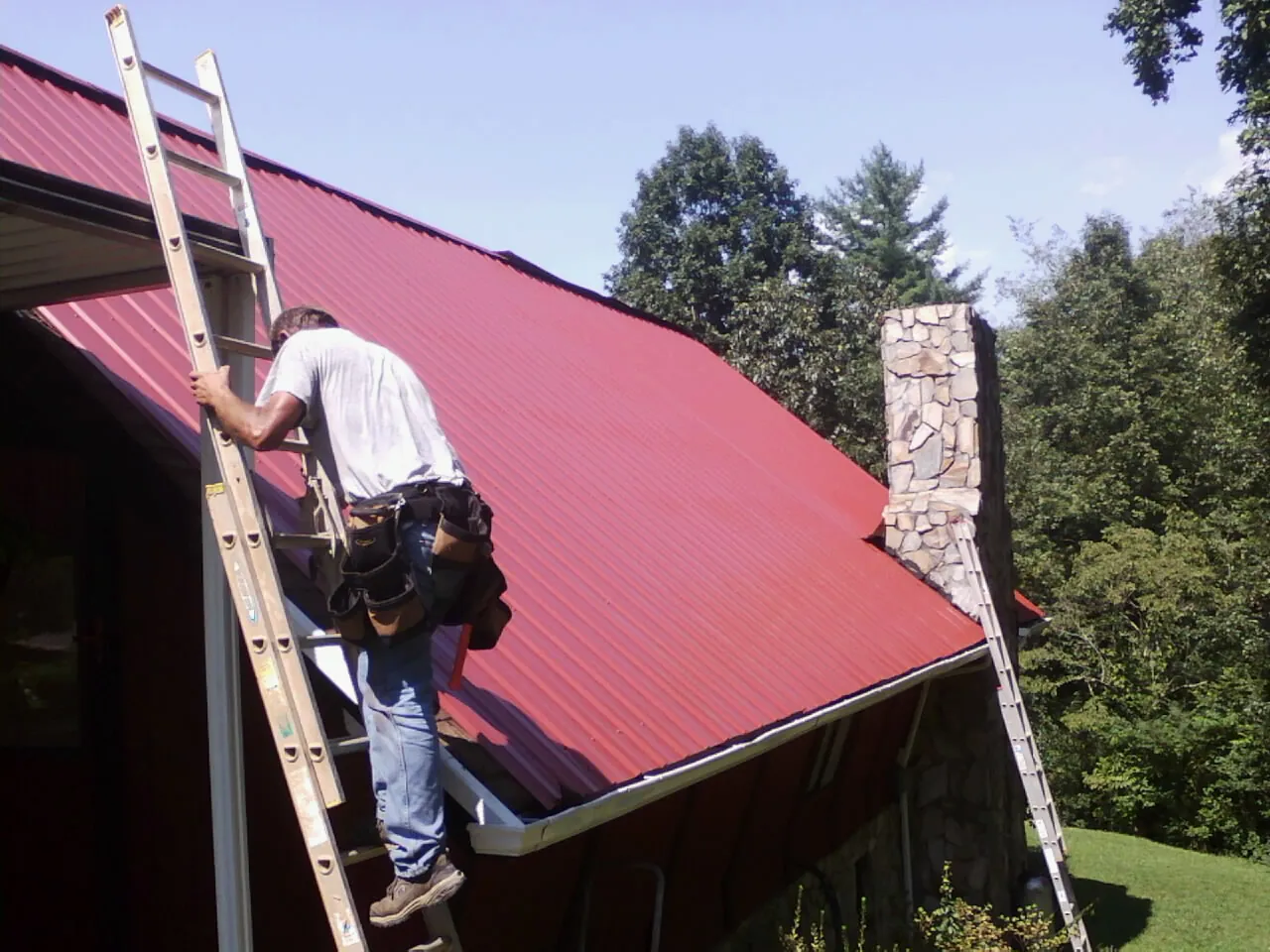 Roofer on a ladder beside a red metal roof and stone chimney — Soffit & Fascia Repair by Roofing Brothers in Lanoka Harbor